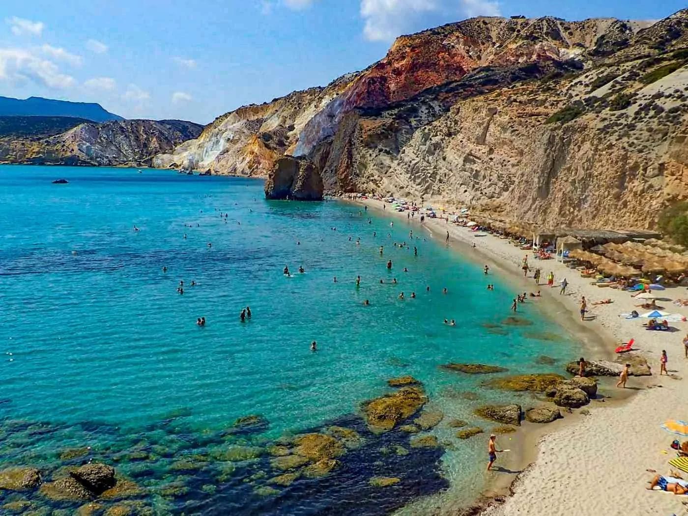 long stretch of sandy beach with straw umbrellas dotting the coastline at Fyriplaka Beach in Milos Greece