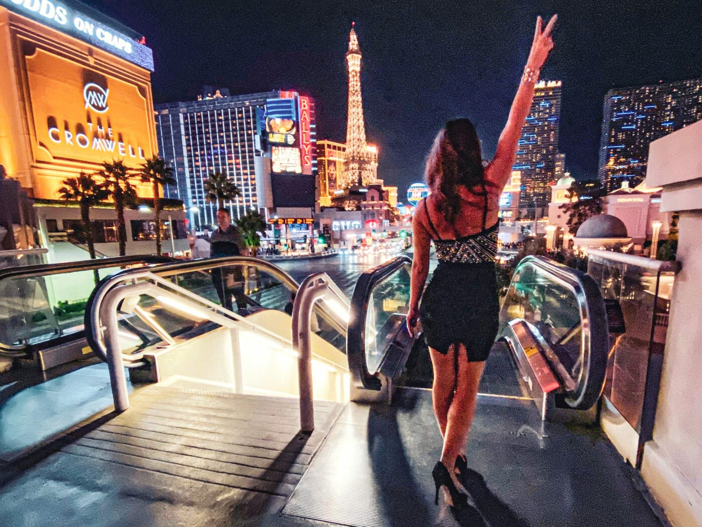 back of a woman dressed in a sparkly dress holding up a peace sign on the Las Vegas strip at night time