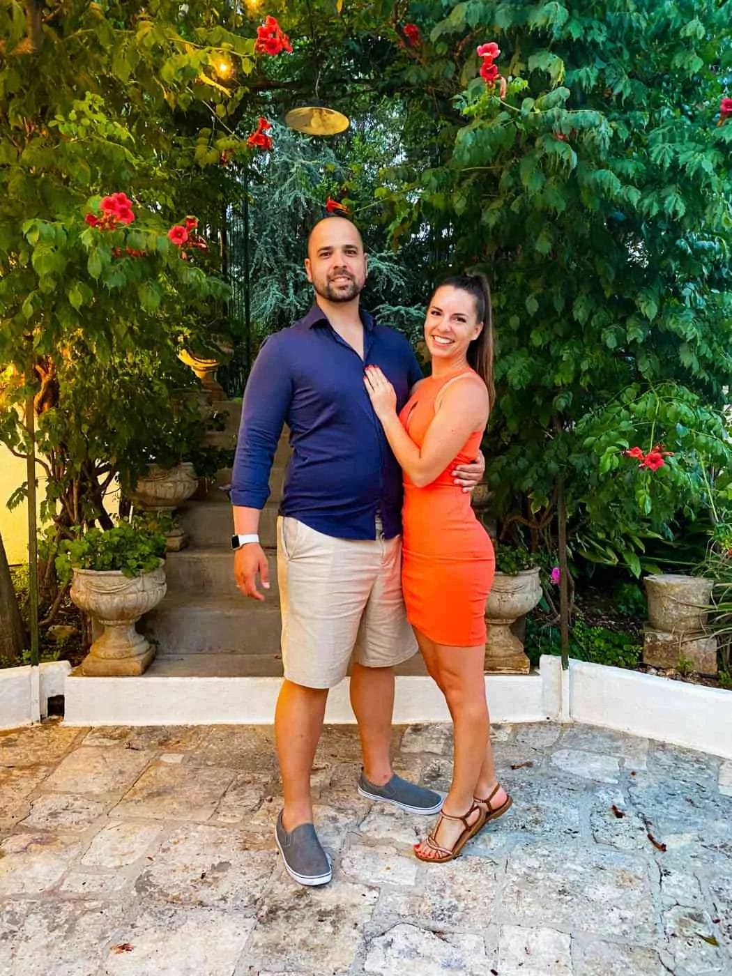 A couple elegantly dressed for summer standing and posing for a picture in front of a flowered bush in the courtyard of Masseria del Frantoio, restaurant in Ostuni Puglia.