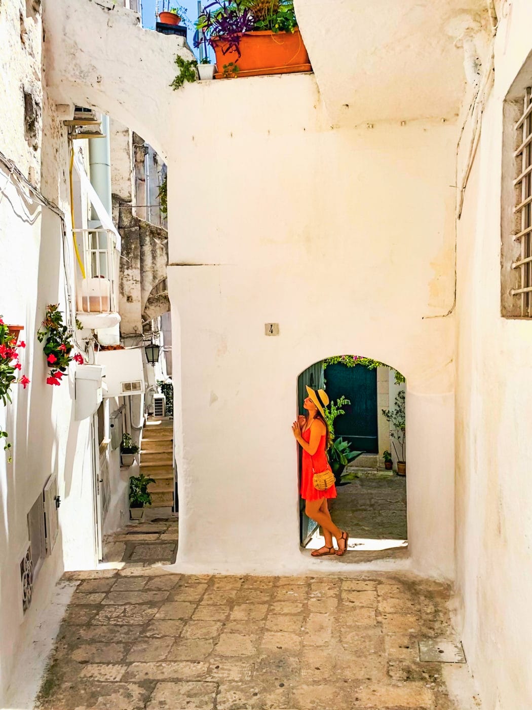 A woman in red dress and straw hat leaning against the archway of a whitewashed building in Ostuni, Puglia Italy
