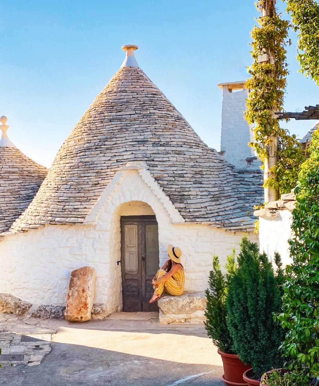 a woman in a yellow dress and hat sitting on the bench outside of a trullo in Alberobello, southern Puglia