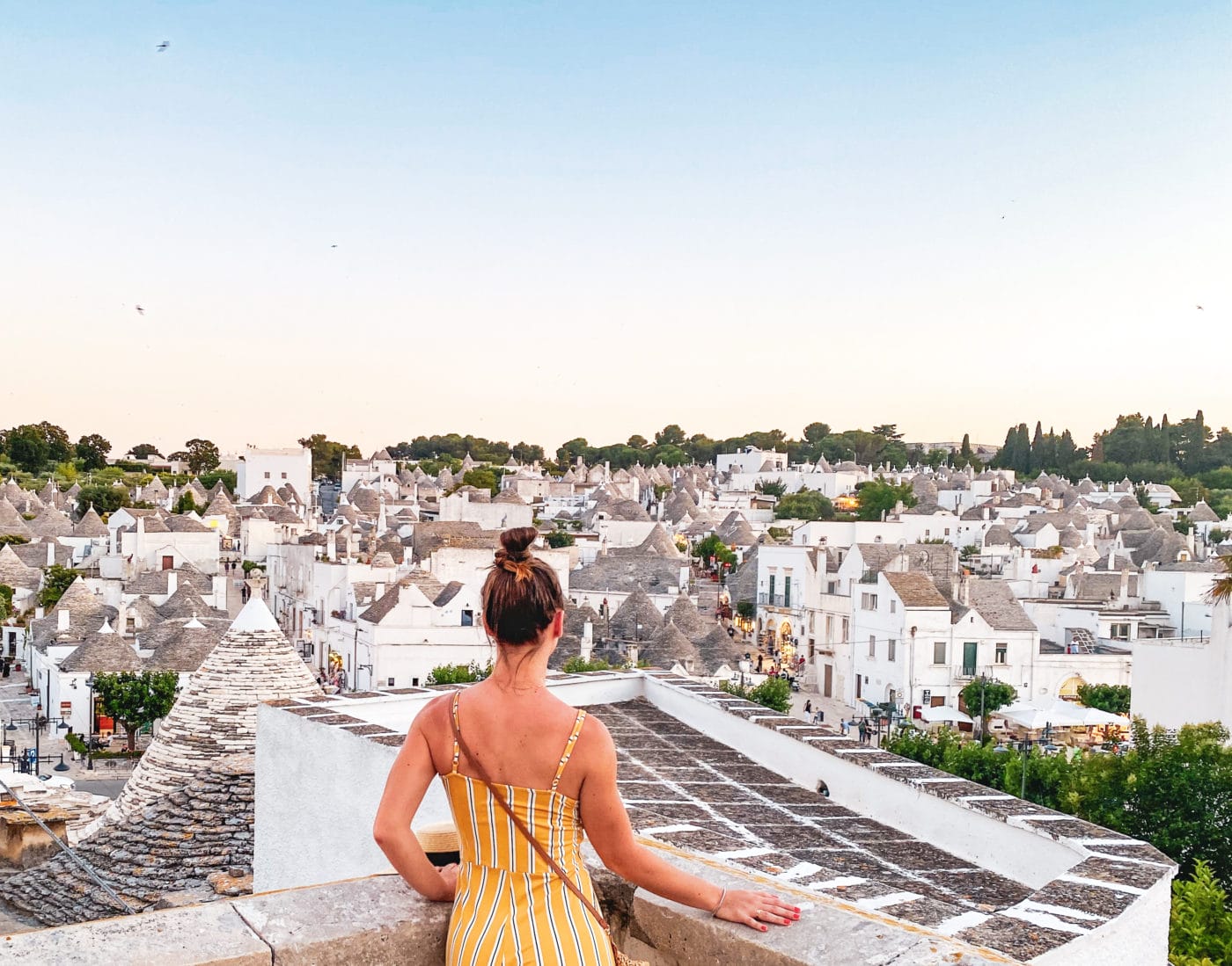 woman in yellow dress and bun looking out from a terrace onto the many trulli below in Alberobello Puglia, Italy
