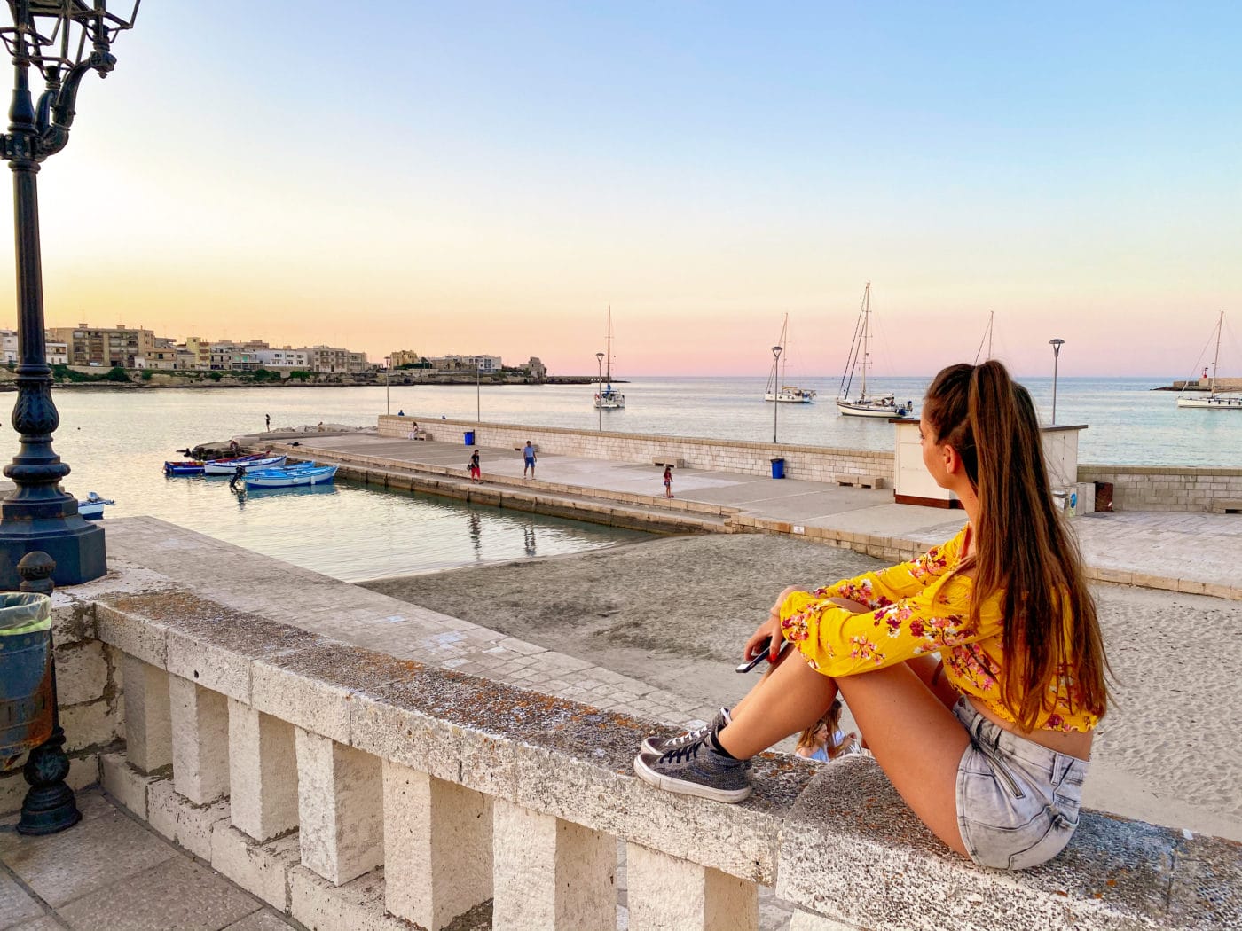 Girl sitting on a low wall on the lungomare degli Eroi, Otranto Puglia staring out at the port and horizon