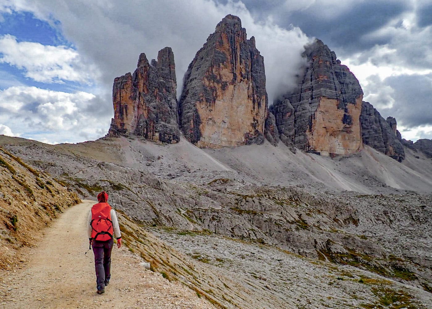 woman with orange backpack hiking away from the camera towards the 3 peaks (tre cime di lavaredo) in the Dolomites, Italy