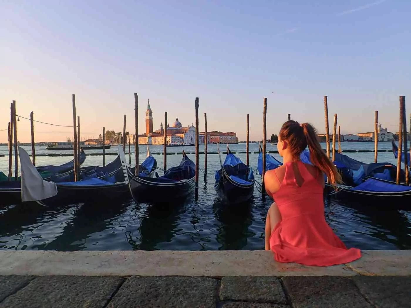 Woman in red dress sitting on the water edge looking at san giorgio island and the many gondolas in venice