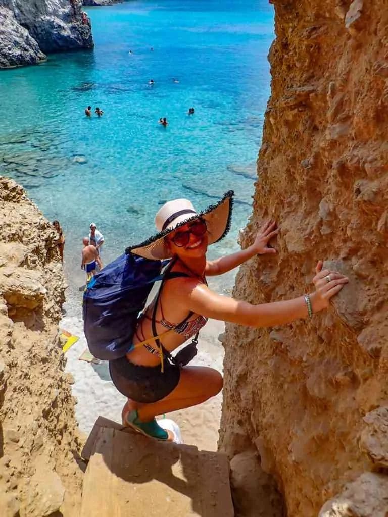 woman smiling for the camera about to climb down a cliff that soars over a beautiful beach with crystalline waters in Milos, Greece.