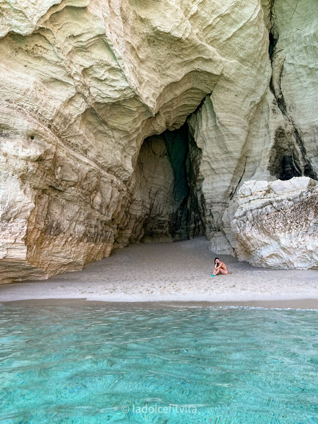 a woman sitting on a beach beneath a grotto in tropea, calabria