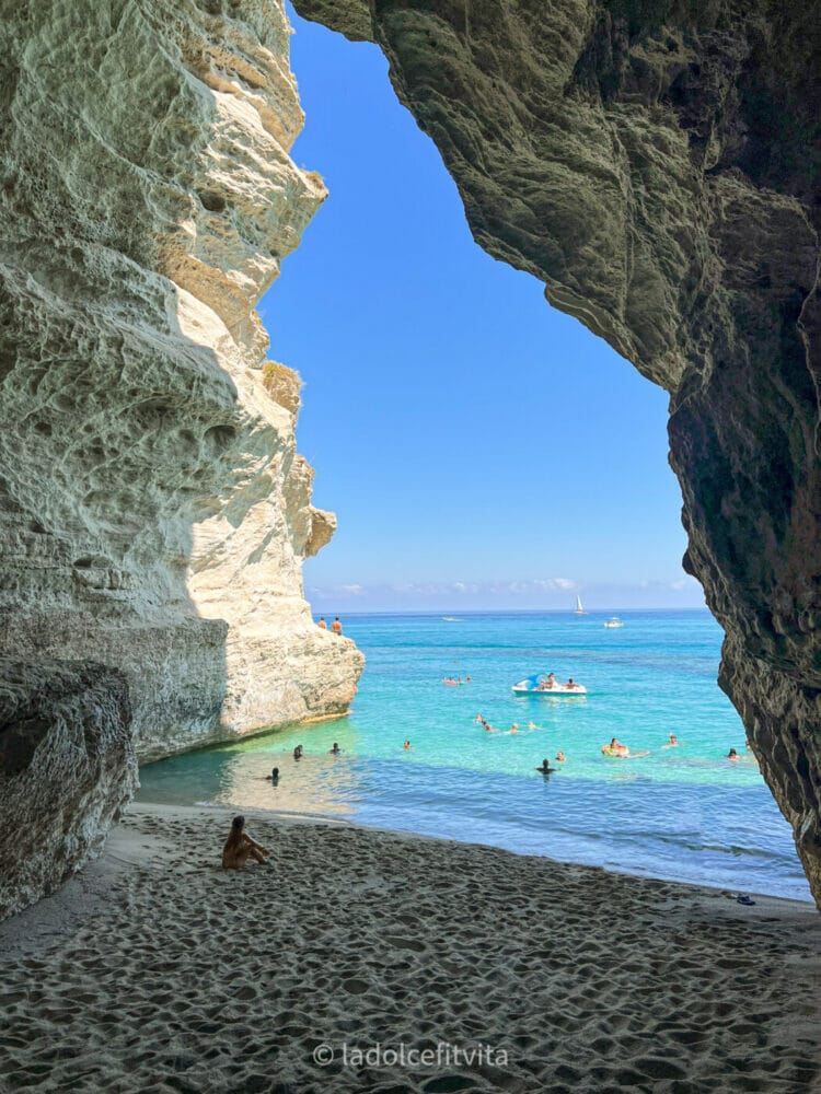 a woman sitting in sand underneath a grotto looking out at turquoise sea waters in Tropea calabria italy