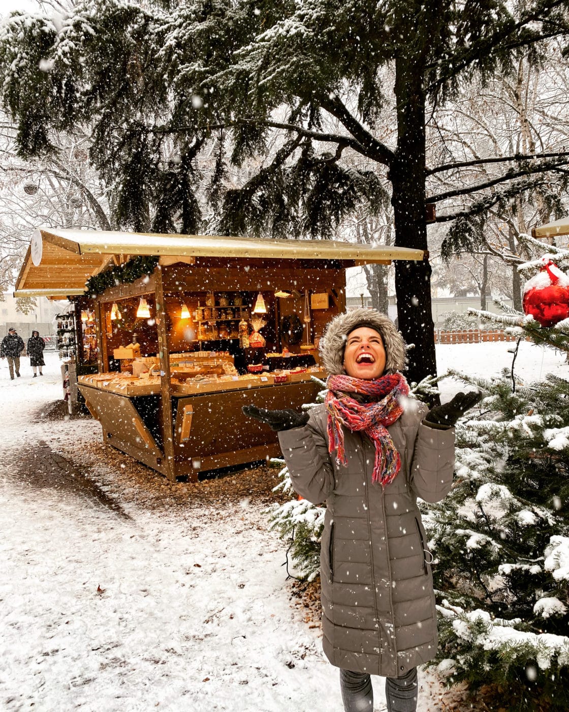 a woman laughing and holding out her hands to the falling slow with a christmas market stall in the background