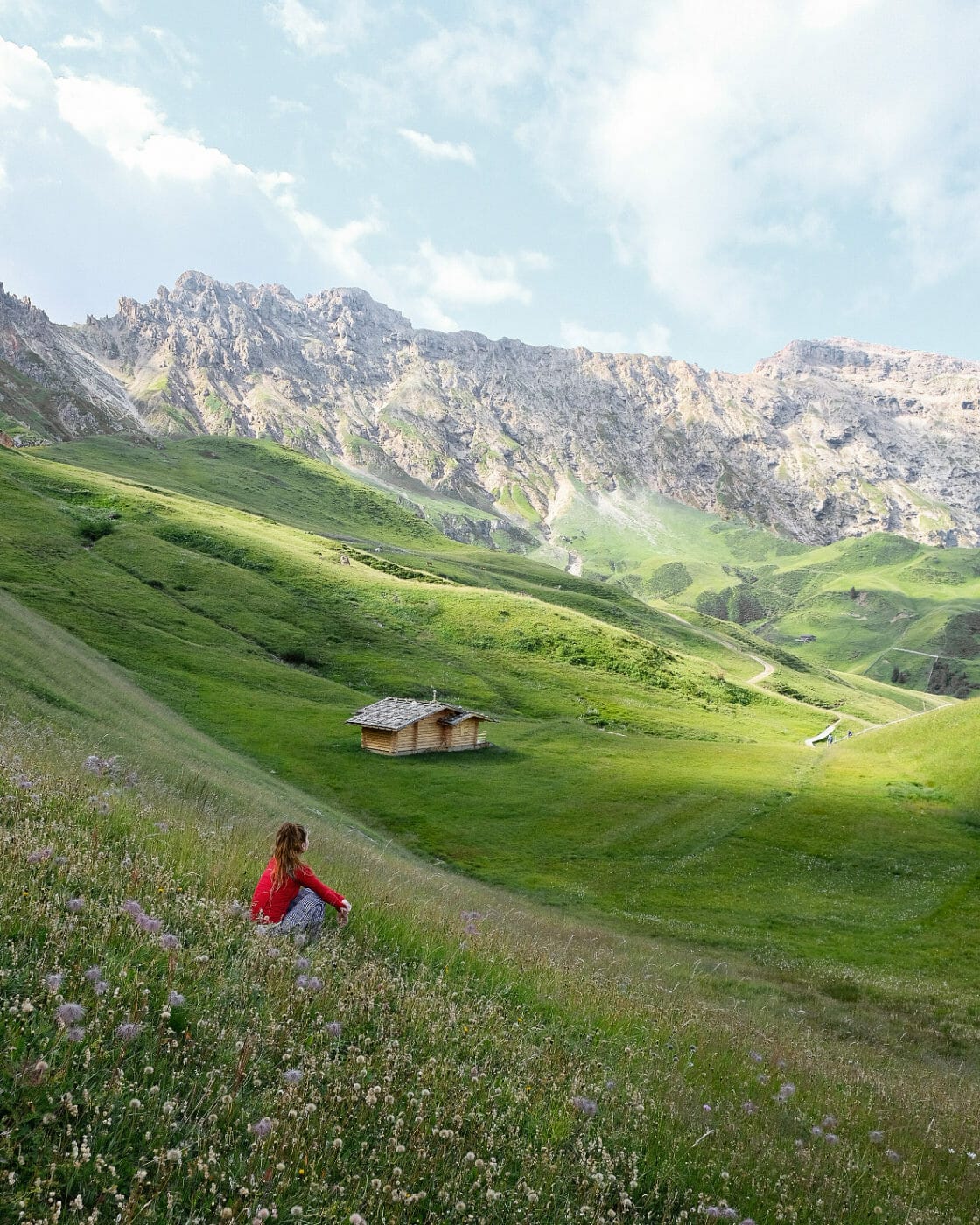Woman sitting in the grass of a meadow gazing at the mountain hut in the distance in the Alpe di Siusi Dolomites