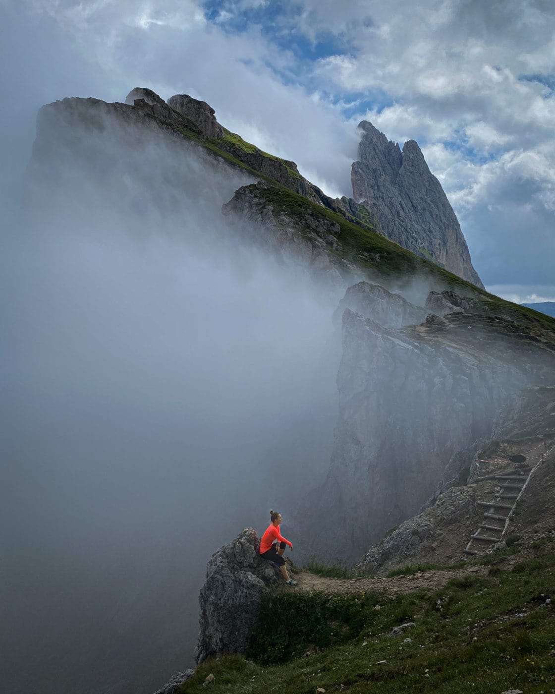 woman hiker amidst fog sitting on the edge of the seceda ridgeline in the dolomites