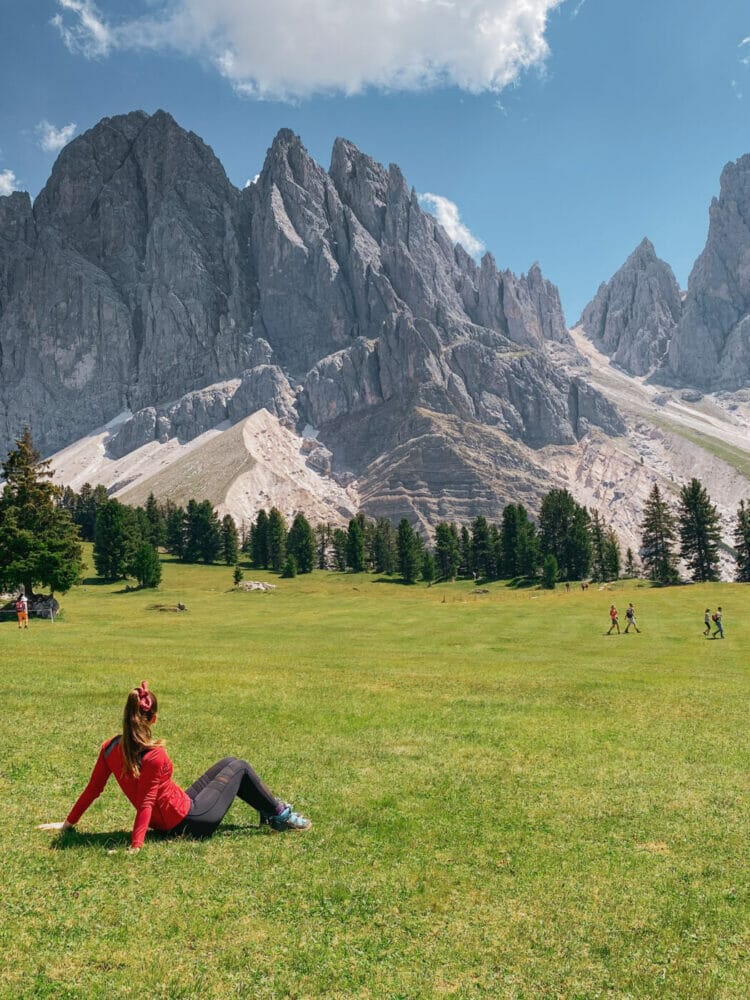 woman in red shirt and black leggings with a ponytail sitting in a wide open meadow gazing at the Odle mountain peaks in the background in the Italian Dolomites