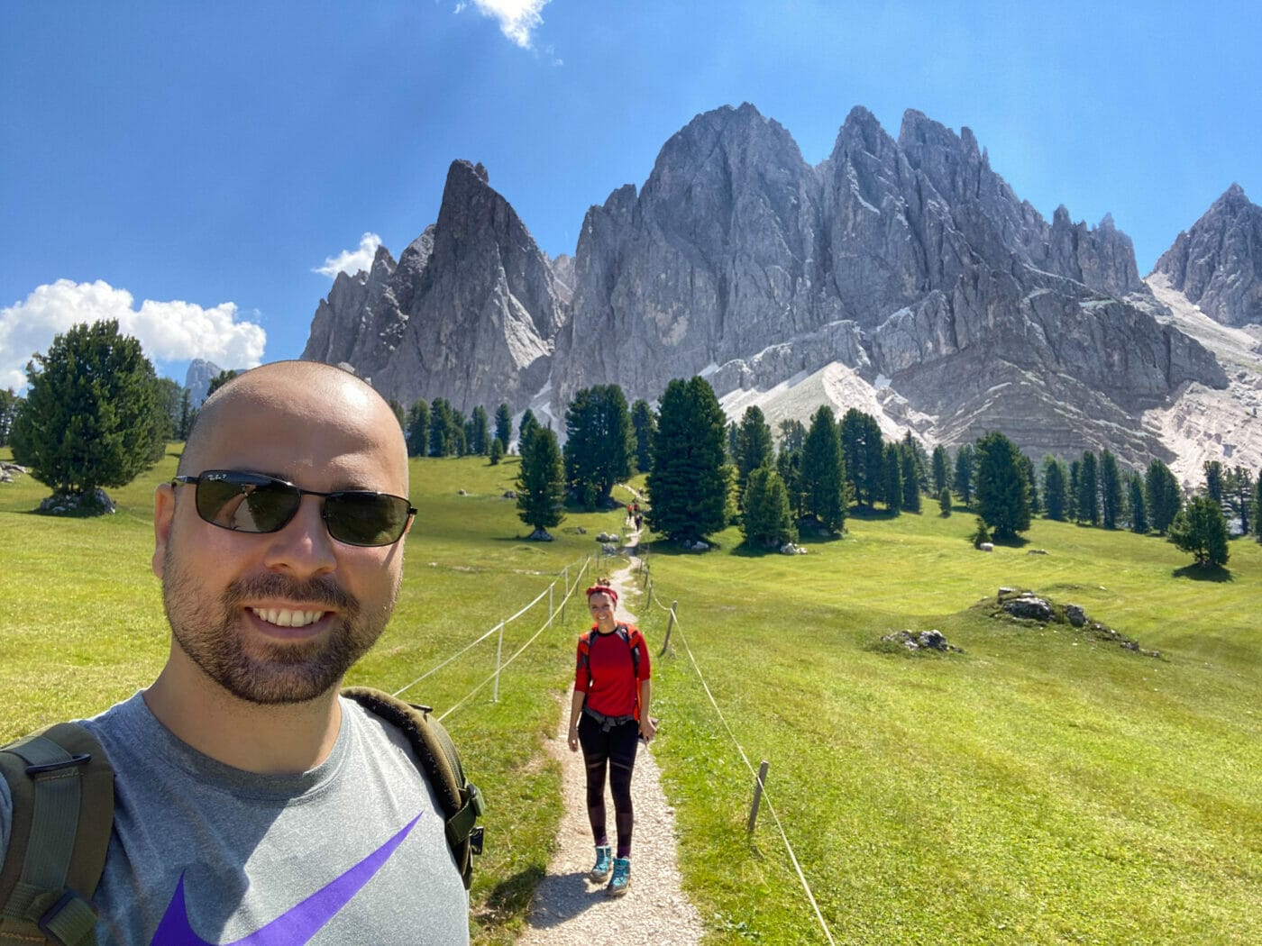 Two hikers posing for a selfie on a footpath with the glorious Odle mountains in the background in the Italian Dolomites.