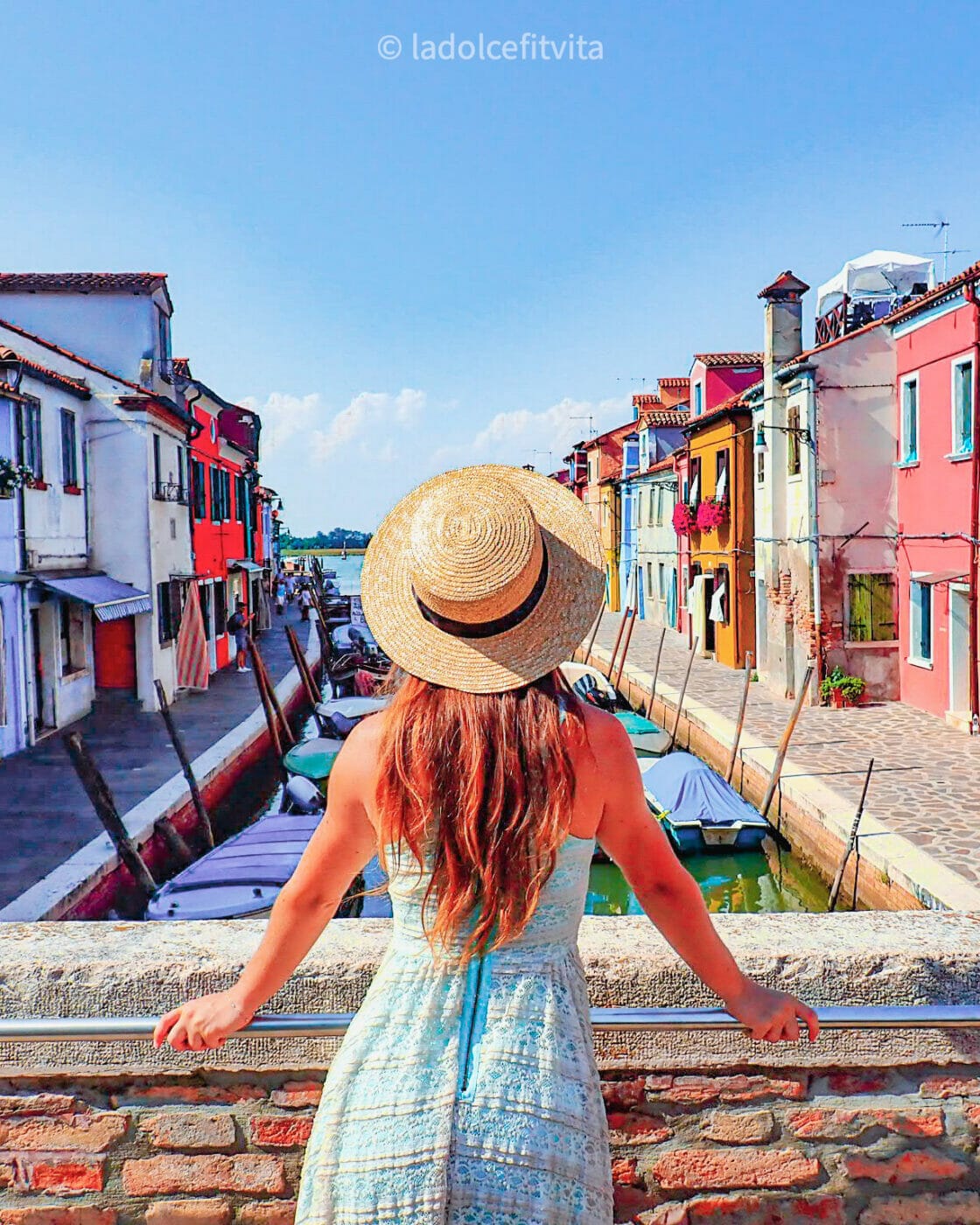 a woman in a summer dress and wearing a sunhat stands on the middle of a bridge stretching over a canal in the very colorful island of Burano in Venice Italy