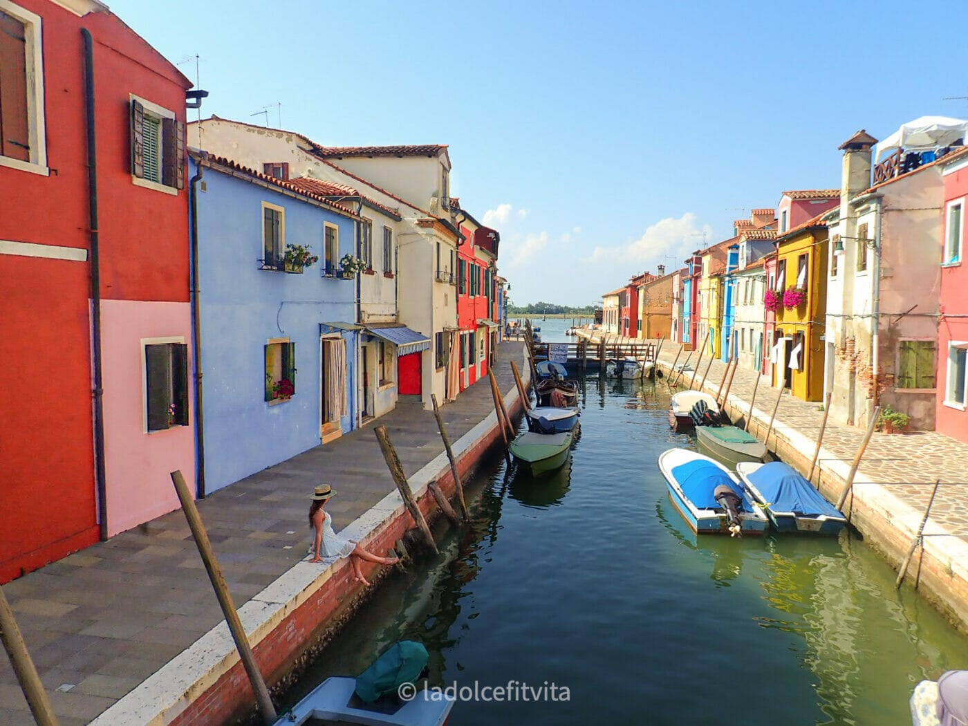 a woman with a hat sitting on the banks of a canal with colorful buildings on the island of burano in venice