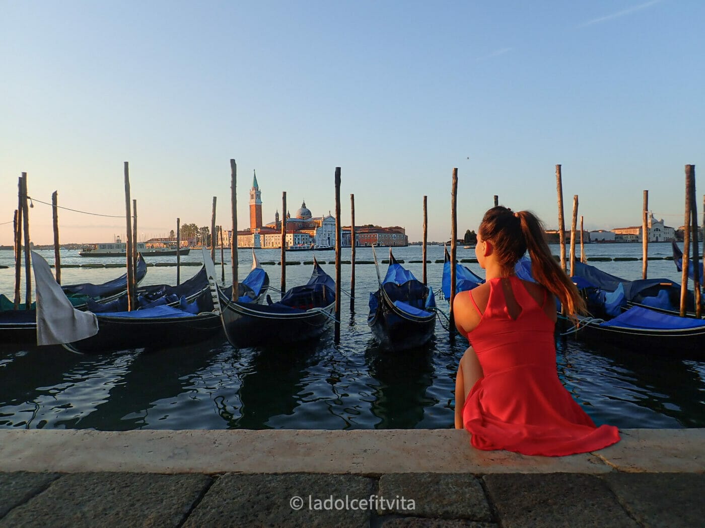 a woman in a red dress sitting on the banks in front of saint mark's square in Venice Italy looking out on san giorgio island