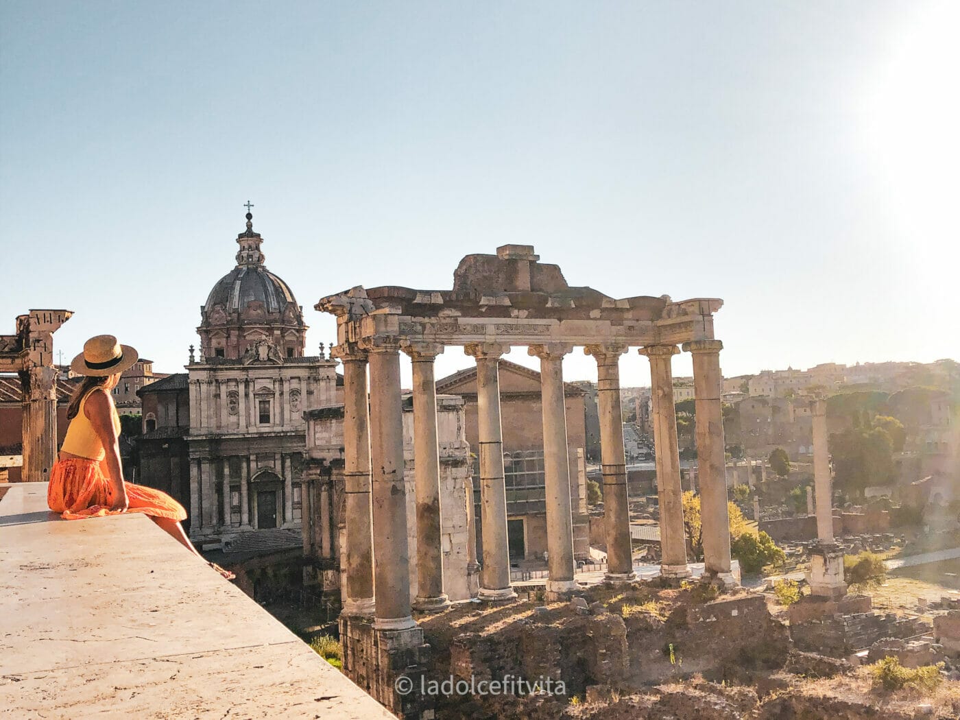 a woman sitting on the edge of wall that overlooks the beautiful Roman forum in Rome Italy at sunset