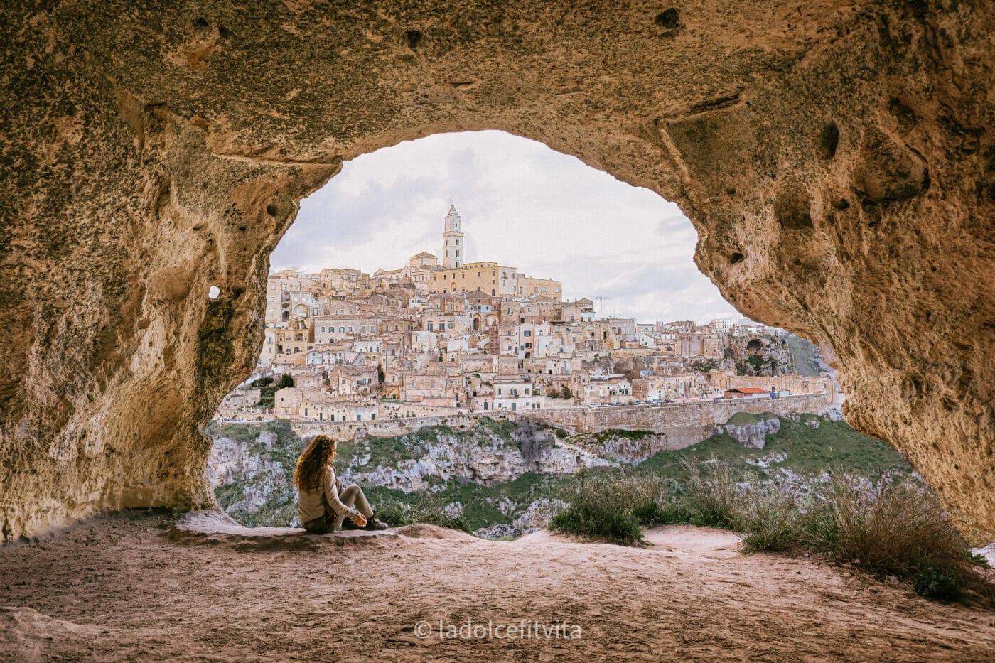 woman sitting down in a cave overlooking the stunning city of matera in italy at sunset