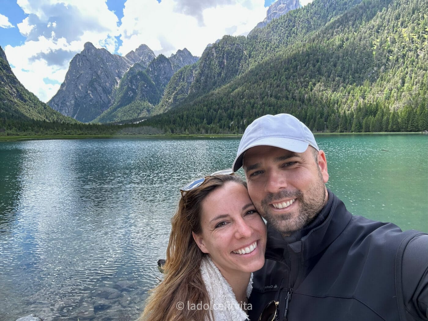 a couple posing for a selfie with mountains and lago di dobbiaco in the background